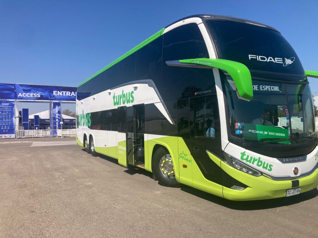 Autobús de Turbus en el aeropuerto de Santiago para el evento FIDAE 2026, rodeado de pasajeros y palmeras.